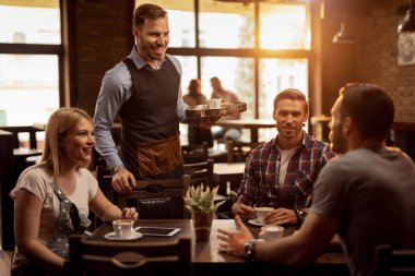 Happy waiter communicating with customers while bringing them their orders in a cafe. 