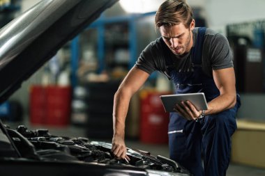 Young auto repairman working on car engine while using digital tablet in repair shop.
