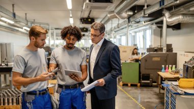 Mid adult manager and two metal workers cooperating while analzying finished steel product and using digital tablet in industrial building. 