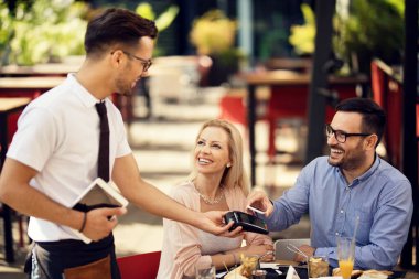 Happy couple using smart phone and paying bill via contactless payment in a bar. 