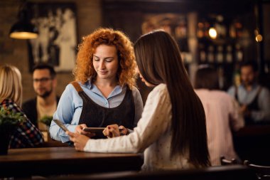 Young smiling waitress writing order on digital tablet while talking to a woman in a pub. 