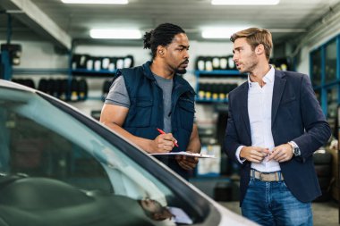 Black car mechanic taking notes while talking to young businessman in auto repair workshop. 