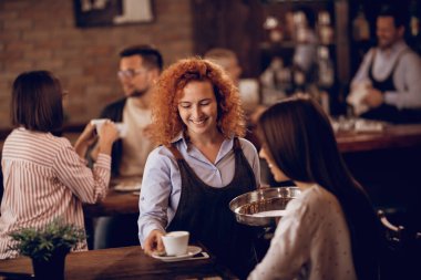 Happy female student serving a guest with a coffee while working as waitress in a cafe. 