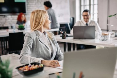 Smiling businesswoman working at her desk and communicating with her colleague who is sitting in the background. 