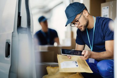 Young manual worker using digital tablet and scanning bar code on label of package in delivery van. 