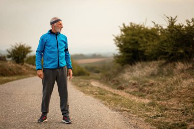 Active mature athlete standing on the road and getting ready for morning run in nature.