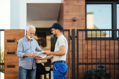 Happy mature man receiving package from a courier and signing paperwork in front of his house. 