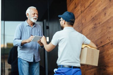 Happy mature man using digital tablet while singing to a postman for home delivery. 