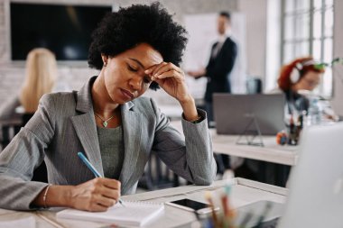 African America female entrepreneur making a plan and writing notes while working at her desk in the office. There are people in the background. 