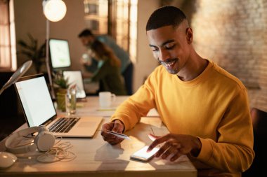 Mutlu Afrikalı Amerikalı girişimci gece çalışırken internet üzerinden yapılan ödemelerden cep telefonu ve kredi kartı kullanıyor.. 