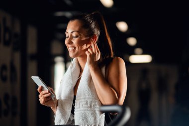Happy athletic woman taking a break after work out and listening music while using smart phone in fitness center. 