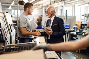 Happy senior CEO shaking hands with young worker while visiting employees at factory plant.