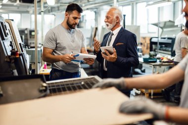 Young worker submit a report to mature businessman while talking in a factory plant. 