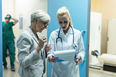 Happy senior woman and female doctor going through medical documents in hospital hallway. 