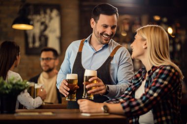 Happy waiter communicating with female guest while serving beer at her table in a bar. 