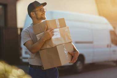 Young delivery man holding stack of packages while making home delivery at residential district. 