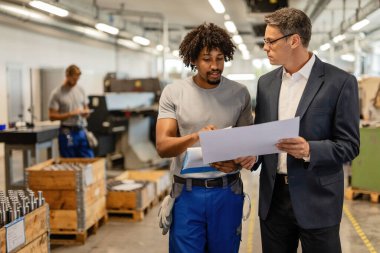 Young black manual worker and quality control manager analyzing production plans and communicating in a factory plant. 