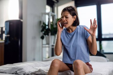Frustrated woman screaming at someone while talking on mobile phone in the bedroom. 