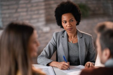 Black female financial consultant taking notes while communicating with her clients during a meeting in the office.
