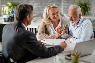 Happy senior woman encourages her husband to sign a contract with their bank manager during a meeting in the office. 