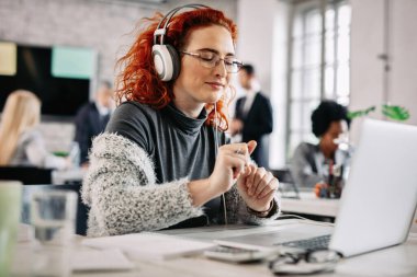 Young happy businesswoman enjoying in favorite music over headphones while working on a computer in the office. There are people in the background. 