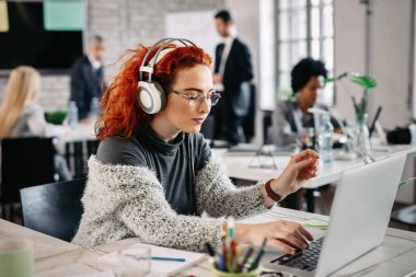 Redhead businesswoman typing an e-mail on laptop while listening music on headphones at work. There are people in the background. 