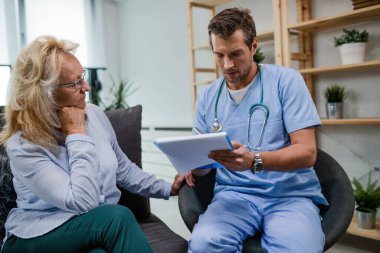 Male doctor looking at medical test results of mature woman while being in a home visit. 
