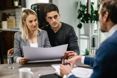 Young couple talking with real estate agent while analzying housing plans during the meeting, 