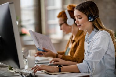 Young businesswoman with headset working on the computer in the office. Her colleague is in the background. 