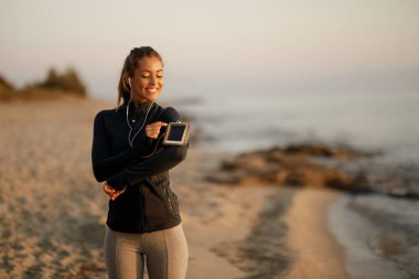 Happy athletic woman preparing for the run and adjusting music on her smart phone at the beach. 