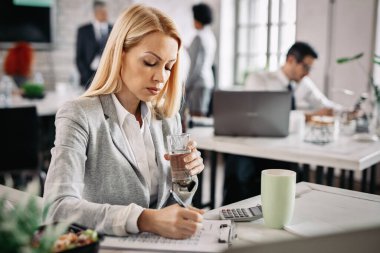 Serious businesswoman writing notes on a document and drinking water in the office. There are people in the background. 