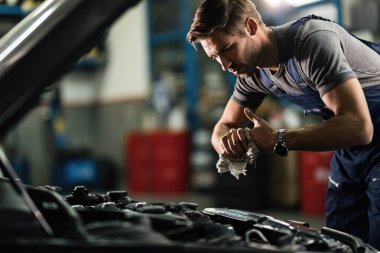 Young mechanic wiping his hands while repairing car engine in auto repair shop. 