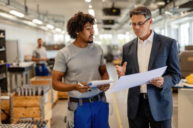Mid adult engineer and African American metal worker cooperating while analzying plans in industrial facility. 