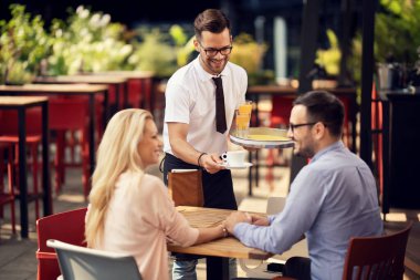 Smiling waiter serving coffee to a couple who is holding hands and talking in a cafe. 