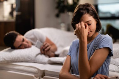 Young woman with headache holding her head in pain while unfaithful boyfriend is texting on mobile phone behind her back.
