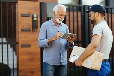 Happy mature man receiving home delivery from a courier and signing on digital tablet. 