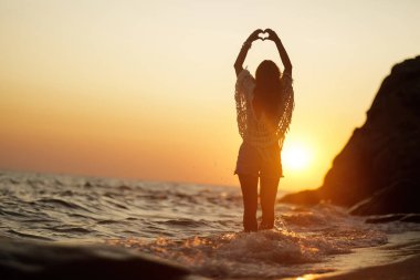 Back view of woman standing in the water and making heart shape during summer sunset. Copy space.