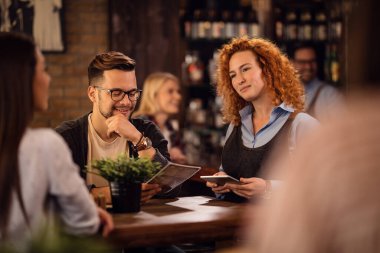 Young smiling waitress using digital tablet while talking to guests and taking their order in a pub. 