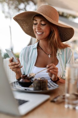 Young smiling woman relaxing in a cafe while eating dessert and typing text message on mobile phone. 