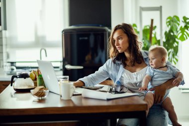 Young working mother using computer while holding her baby and planning home finances. 