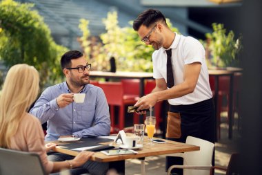 Happy waiter preparing table for dining while his guest are drinking coffee in a restaurant. 