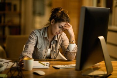 Young displeased woman thinking while working on desktop PC in the evening at home. 