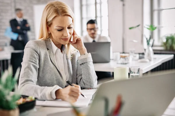 Happy businesswoman working at her desk and taking notes in her note pad. There are people in the background. 
