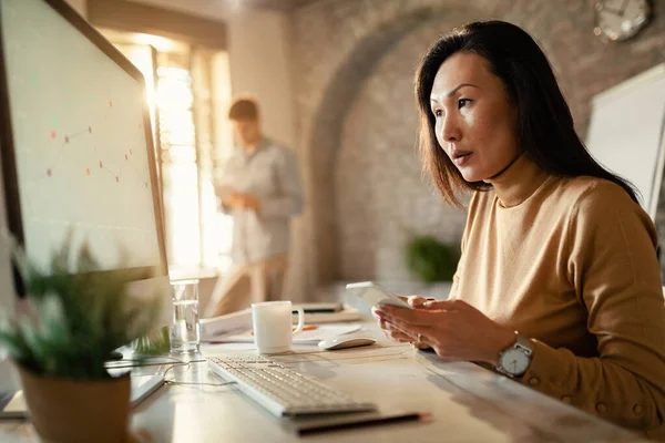 Female Asian entrepreneur reading charts on computer monitor and texting on smart phone in the office.