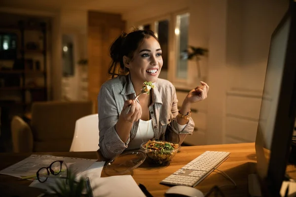 Smiling woman reading an e-mail on desktop PC and eating salad in the evening at home.