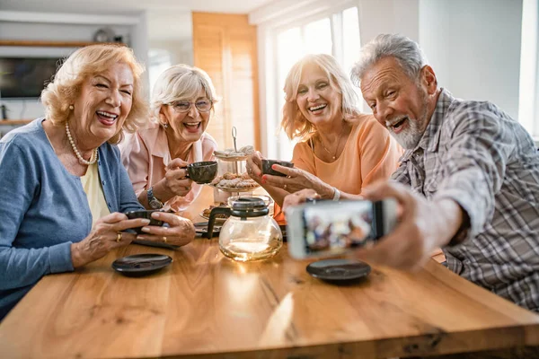 Group of happy seniors drinking tea and having fun while taking selfie with mobile phone at home.