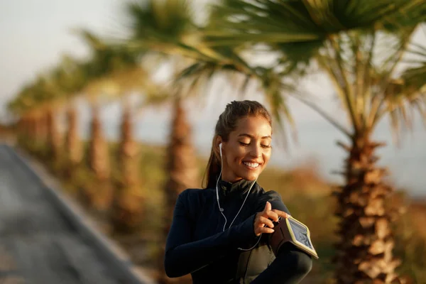 Young athletic woman getting ready for exercising and adjusting mobile phone on arm band in nature.