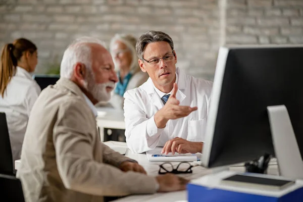 Male doctor explaining to senior patient medical insurance policy while using computer during consultations at clinic.