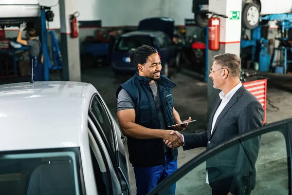 Happy African American car mechanic shaking hands with a customer in auto repair shop.