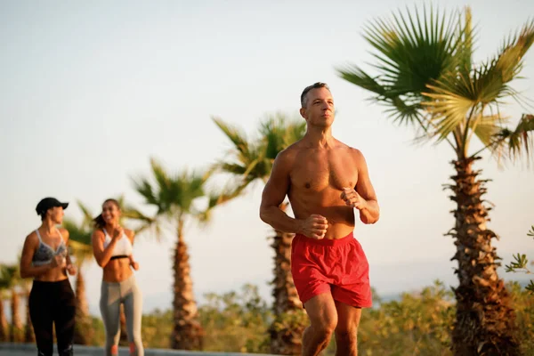Mid adult male athlete running shirtless in nature. Two women are running behind him.